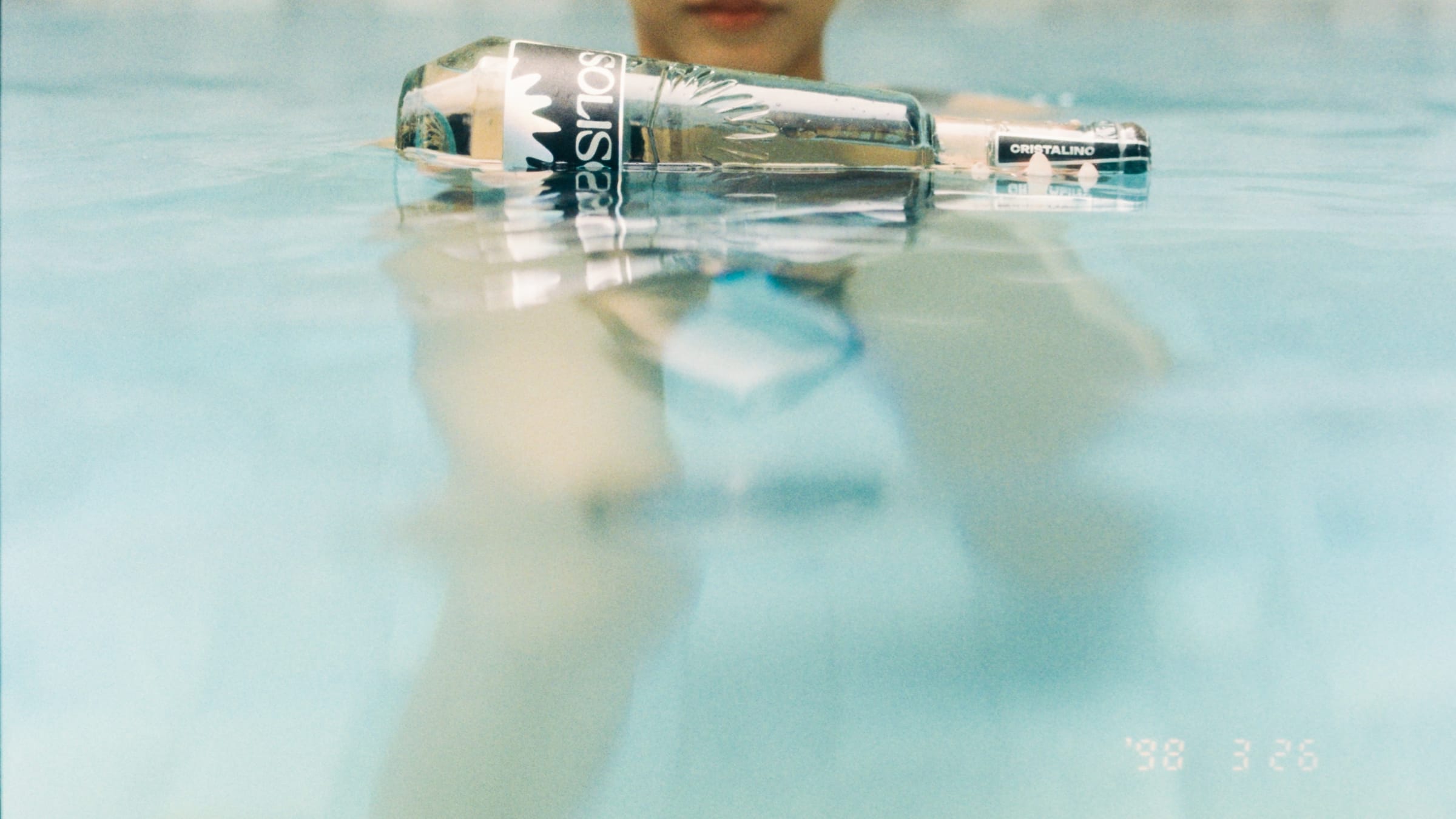 A Solisca bottle floating at the edge of a sunlit pool, a swimmer blurred behind it.