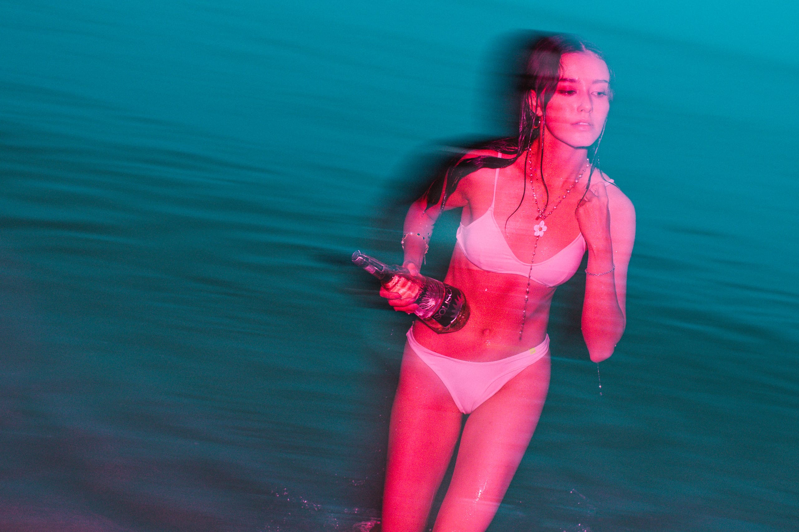A woman wading through dark teal water at night holding a bottle of Solisca, lit in warm pink.