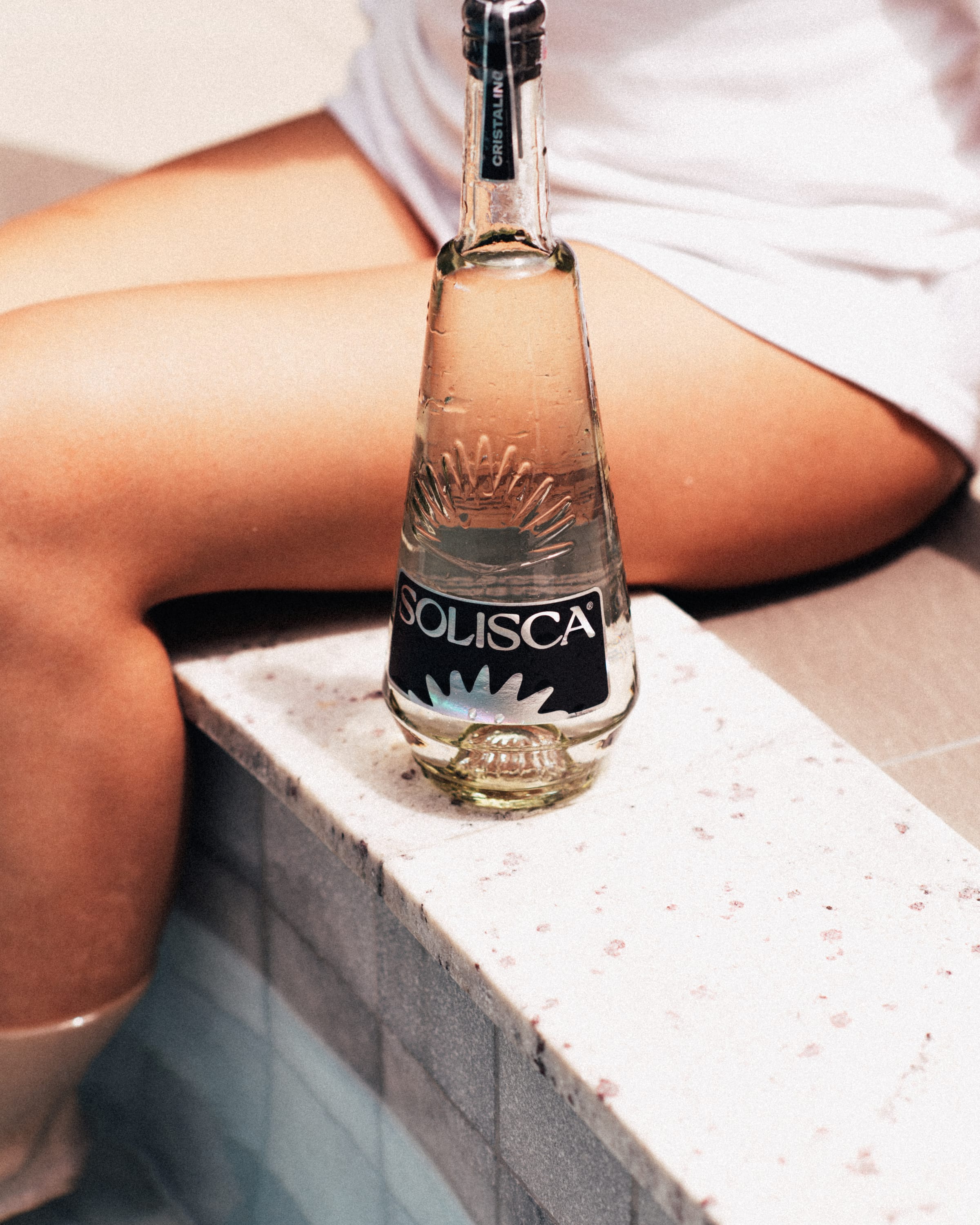 A Solisca Cristalino bottle standing on pool coping beside bare legs in daylight.