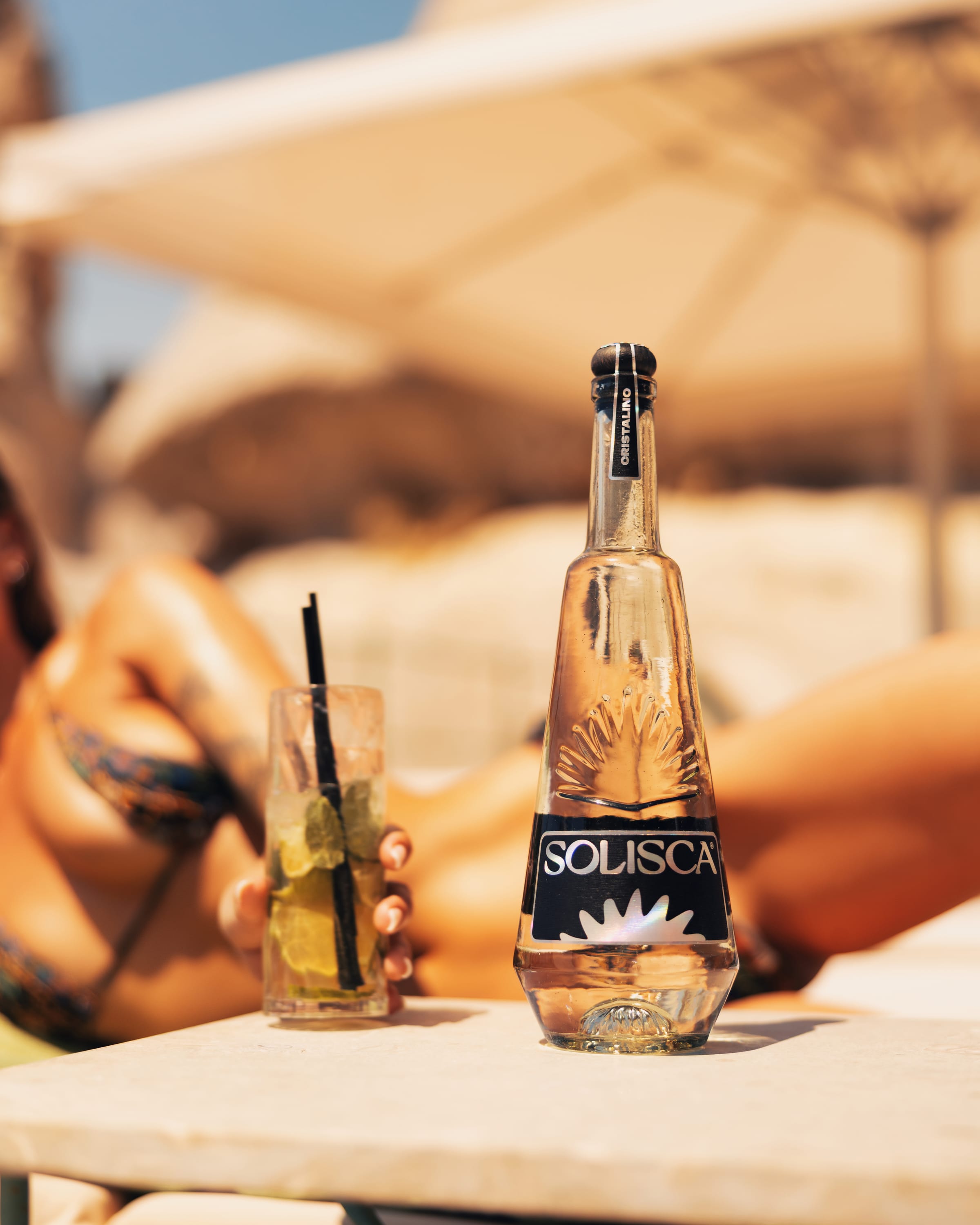 A Solisca Cristalino bottle and a mojito on a sunbed table, tanned skin behind.