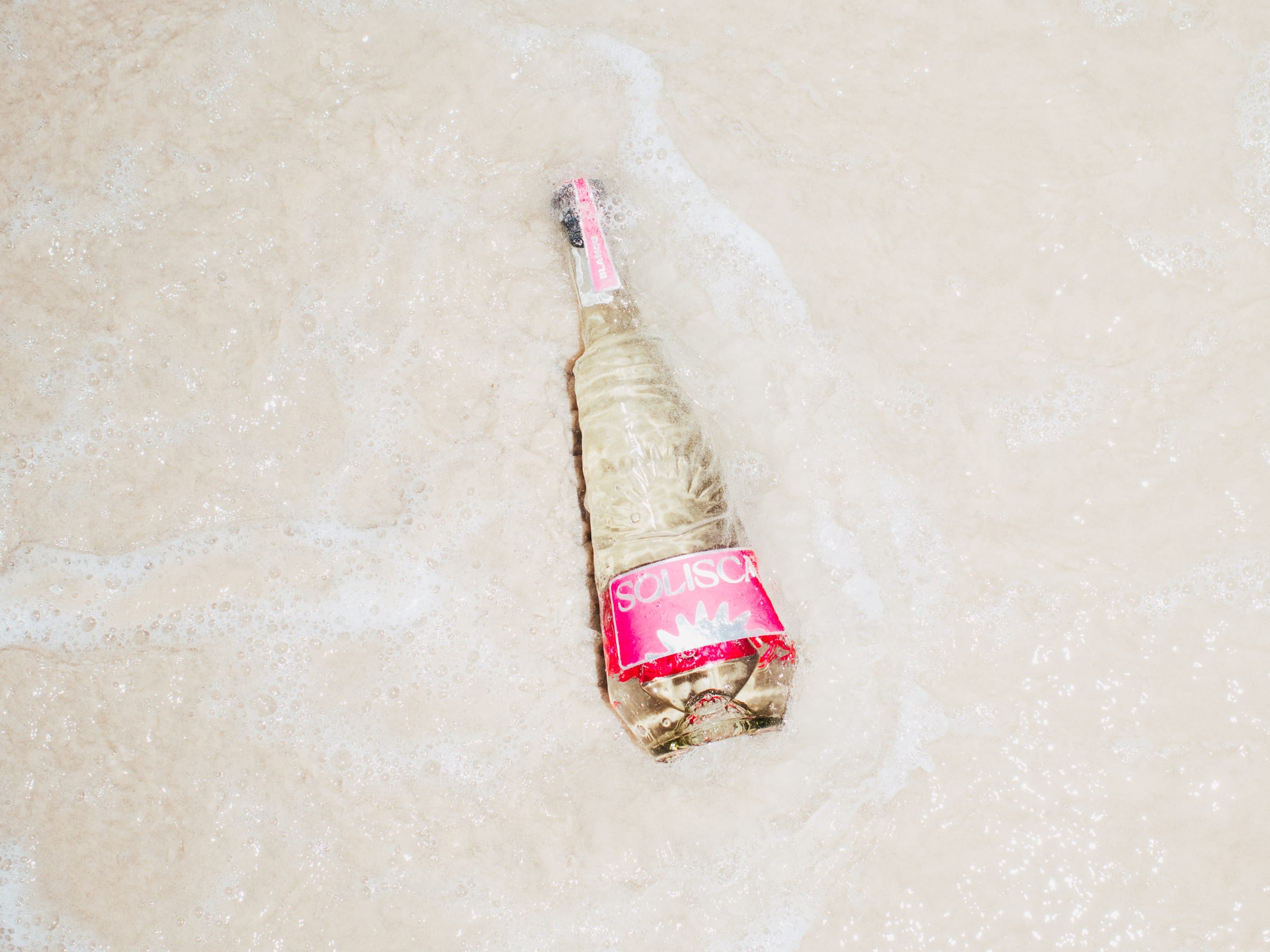 A Solisca Blanco bottle with a pink label lying in white surf foam on the sand, seen from above.