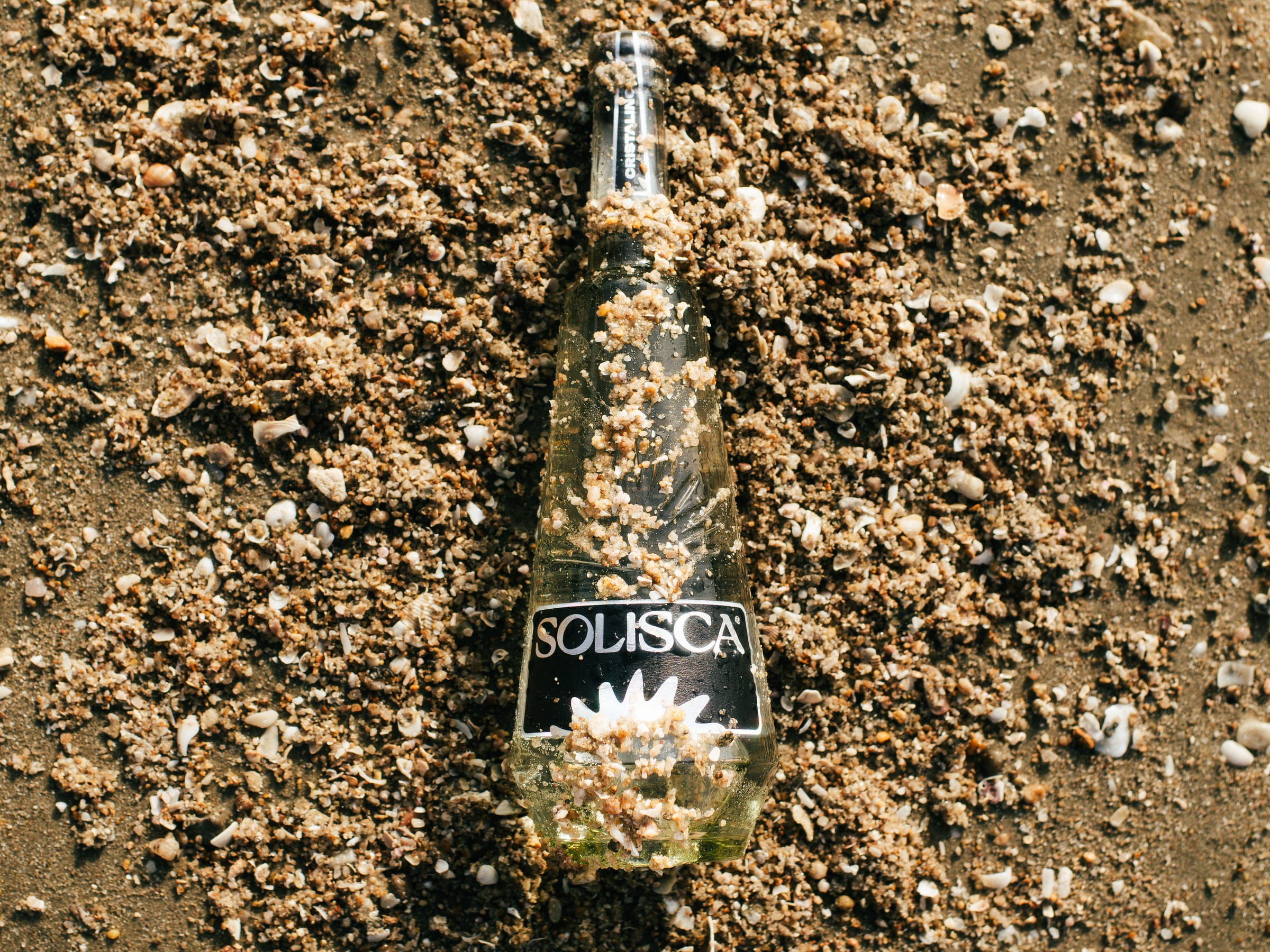 A Solisca Cristalino bottle with a black label bedded in shell gravel on a beach, seen from above.
