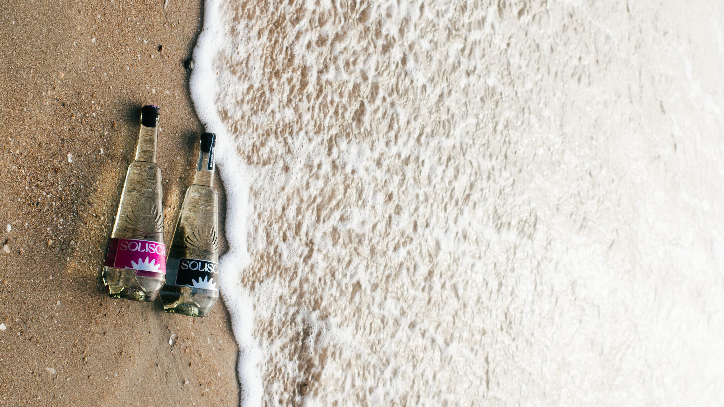 Solisca Blanco and Cristalino bottles lying side by side where surf foam meets the sand.