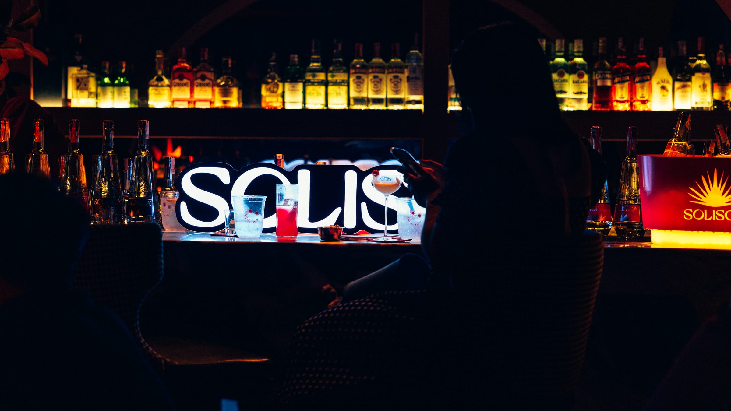 A dark bar at night, a glowing Solisca sign on the counter, backlit bottles on the shelves.